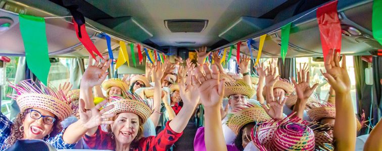 Pessoas vestidas com trajes juninos levantam as mãos dentro de um ônibus. O lugar está com bandeirolas.