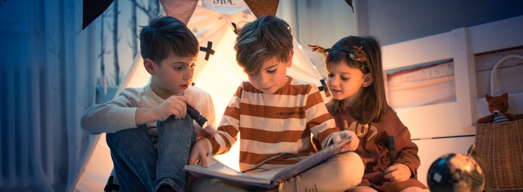 Três crianças estão reunidas, lendo um livro, que está no colo da criança do meio. Elas estão se divertindo nas férias de julho.